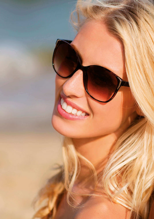 A woman with long blonde hair, wearing stylish sunglasses and smiling brightly at the beach.