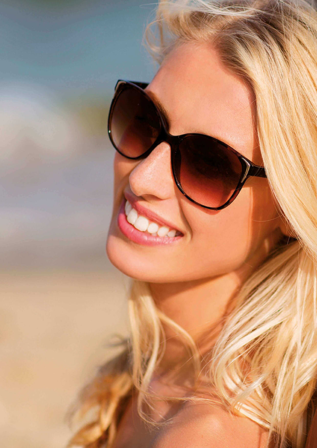 A woman with long blonde hair, wearing stylish sunglasses and smiling brightly at the beach.